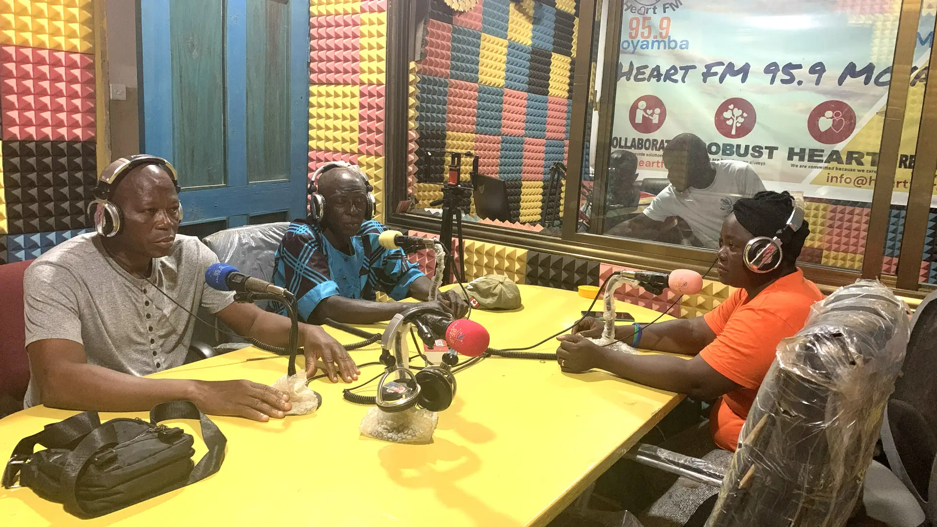 Four Sierra Leonian people sit in a radio studio They are all wearing headphones and speaking into microphones. One is behind a screen on a desk