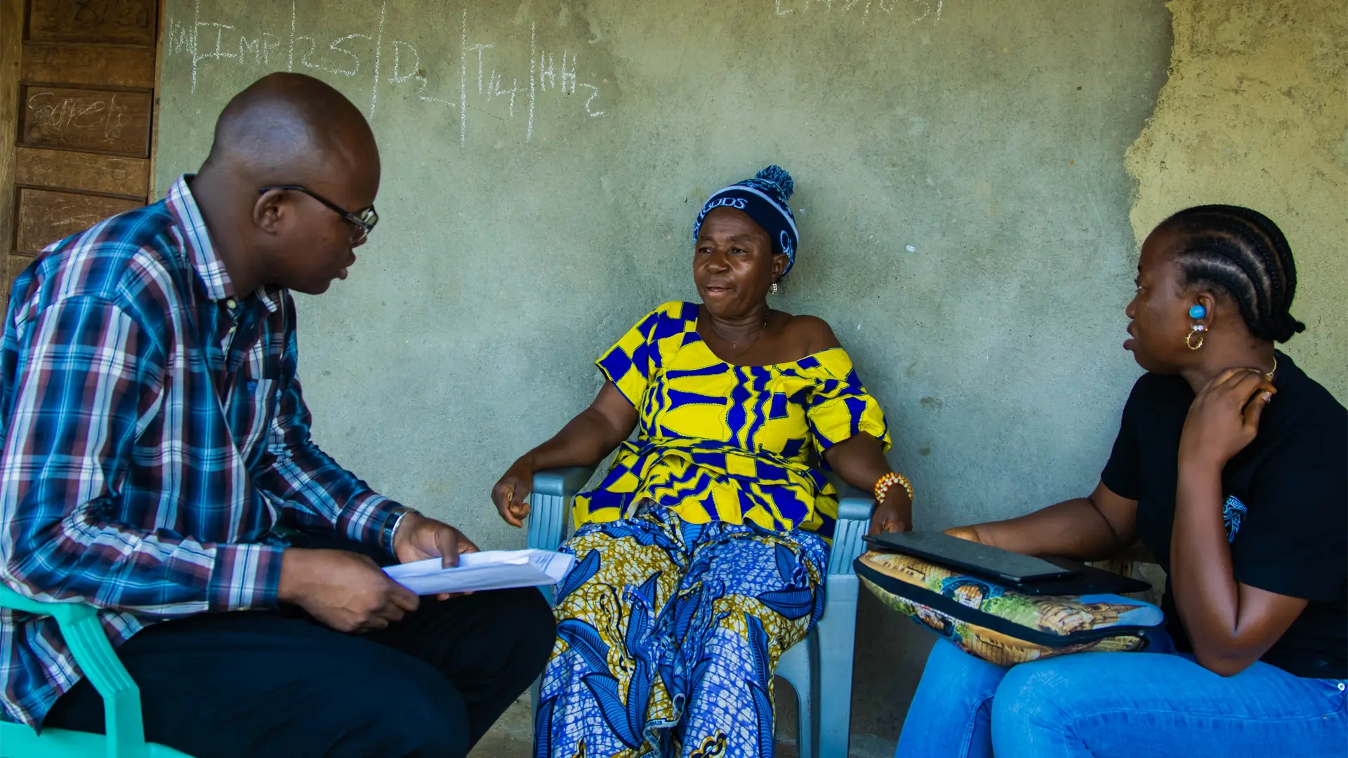It's a verandah in Sierra Leone. A young woman and a man with papers and laptop speak to an older woman wearing a bobble hat